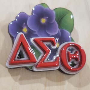 Könnte beinhalten: Dekorative Anstecknadel mit lila Blumen und roten griechischen Buchstaben: Delta, Sigma und Theta. Die Anstecknadel hat einen weißen Rand und ist vor einem hellen Holzgrund platziert. Die Blumen haben gelbe Zentren und grüne Blätter.