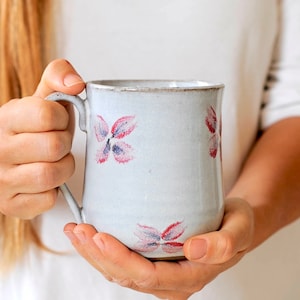 May include: A white ceramic mug with a handle, featuring a floral design in shades of pink and purple. The mug is slightly curved and has a textured surface.