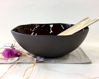 May include: A dark gray ceramic bowl with a glossy, dark interior. Two light-colored wooden serving spoons rest inside. The bowl sits on a gray linen cloth, with purple flowers and dried grass accents.