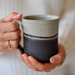 Peut inclure: Une tasse en céramique avec un glaçage noir et blanc. La tasse a une large anse arrondie et une lèvre légèrement évasée.