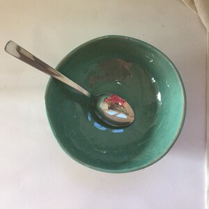 Op de afbeelding: Een teal keramische kom met een zilveren lepel erin. De kom heeft een licht getextureerd oppervlak.