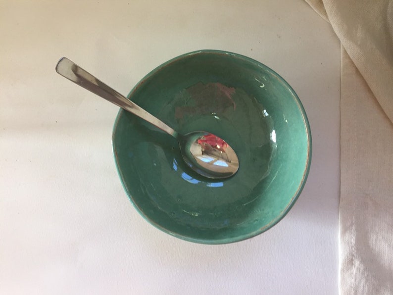 Op de afbeelding: Een teal keramische kom met een zilveren lepel erin. De kom heeft een licht getextureerd oppervlak.