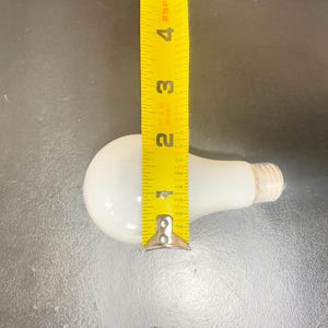 May include: A white light bulb with a yellow measuring tape next to it. The bulb is approximately 4 inches long. The base of the bulb is a light tan color. The background is a dark gray surface.