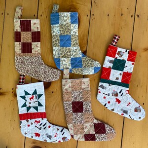 May include: Five quilted Christmas stockings with different patterns. The stockings are made of various fabrics, including red, green, blue, and brown. The stockings are hanging on a wooden surface.