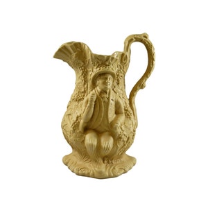 May include: A cream-coloured ceramic jug with an ornate design featuring a man in a hat and a vine motif.