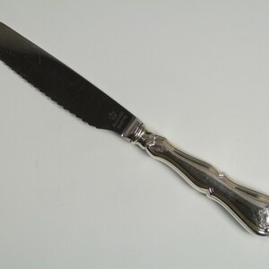 May include: A silver serrated butter knife with a decorative handle. The knife has the words "Wallace Sterling" engraved on the blade.