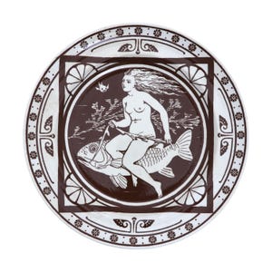May include: A round, white ceramic plate with a brown illustration. The illustration shows a nude figure riding a fish, with a bird in the background. The plate has a decorative border with floral and geometric patterns.