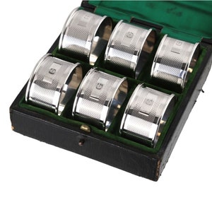 Könnte beinhalten: Sechs silberne Serviettenringe mit einem strukturierten Design und dem Buchstaben "G" auf jedem Ring graviert. Die Ringe befinden sich in einer schwarzen Box mit grünem Samtbezug.