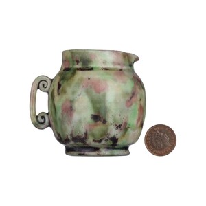 May include: A small green ceramic jug with a mottled glaze. The jug has a rounded body and a single handle with a scroll design. The jug is about the size of a UK penny.