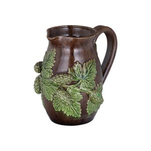 May include: A brown ceramic pitcher with a green and brown leafy design. The pitcher has a handle and a rounded body.