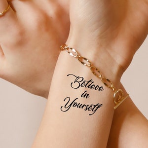 Könnte beinhalten: Ein temporäres Tattoo auf dem Handgelenk einer Person, auf dem in kursiver Schrift "Glaube an dich selbst" steht.
