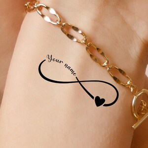 Puede incluir: Un tatuaje de un símbolo de infinito negro en la muñeca de una persona con las palabras "Your name" escritas dentro del símbolo. Un pequeño corazón negro se encuentra en la parte inferior del símbolo de infinito. La persona lleva una pulsera de cadena dorada.