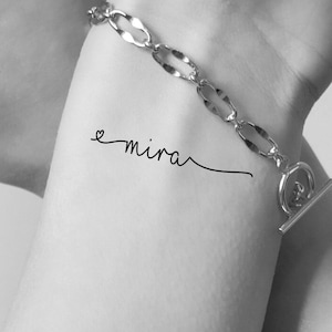 May include: A black and white photo of a person's wrist with a small tattoo that says "e mira" in cursive script. The person is wearing a silver chain bracelet.