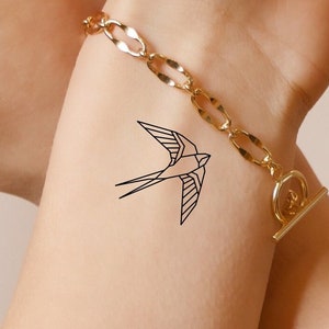 Pode incluir: Um desenho de linha simples de uma tatuagem de andorinha no pulso de uma pessoa. A tatuagem é preta e está logo acima de uma pulseira de corrente dourada.