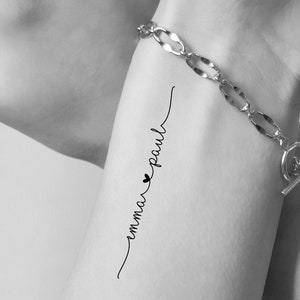 May include: Black and white image of a wrist adorned with a silver bracelet and a delicate ring. A temporary tattoo on the wrist reads "emma ♥ paul" in cursive script, with a small heart symbol.