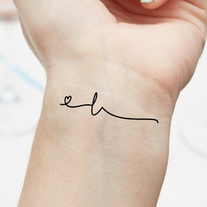 Peut inclure: Un tatouage temporaire simple sur le poignet d'une personne qui dit "eh" avec un petit cœur devant le "e".