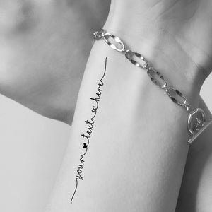 Puede incluir: Una pulsera de cadena plateada con eslabones ovalados y un cierre de barra. La pulsera se muestra junto a un tatuaje temporal en la piel que presenta el texto en cursiva negra "your text here" con un pequeño icono de corazón.