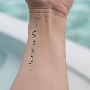 Puede incluir: Primer plano de la muñeca de una persona con un simple tatuaje negro que dice "hudson charlotte" en cursiva.