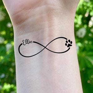 Puede incluir: Tatuaje negro con el símbolo del infinito con el nombre "Ellie" en escritura cursiva a la izquierda y una huella de pata a la derecha. El tatuaje está sobre piel clara.