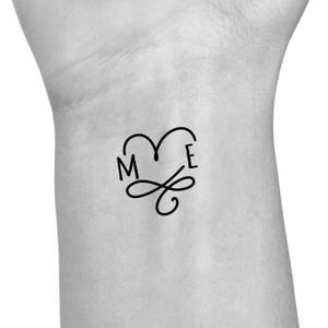 May include: A black temporary tattoo design featuring the letters "M" and "E" within a heart shape, with an infinity symbol below. The tattoo is applied to the skin.