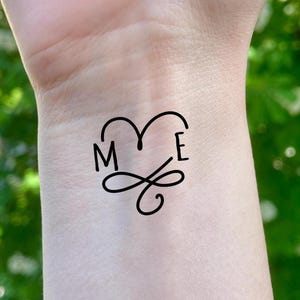 May include: Black temporary tattoo with the letters "M" and "E" inside a heart shape, with a decorative flourish below. The tattoo is on a person's wrist, with a blurred green background.