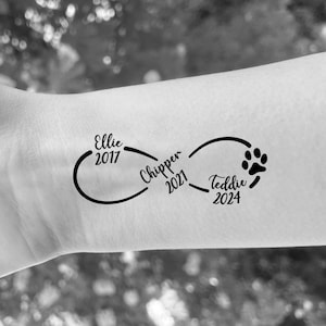 May include: Black infinity symbol tattoo with the names "Ellie 2017", "Chipper 2021", and "Teddie 2024" along with a paw print. The tattoo is on a light skin tone.