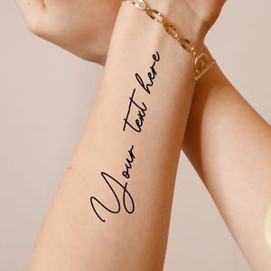 May include: A close-up of a person's arm with a gold bracelet. The arm has a temporary tattoo with the words "Your text here" in black cursive script. The skin tone is light.