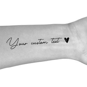 May include: Temporary tattoo featuring the words "Your custom text" in a handwritten style, accompanied by a small black heart. The design is applied to the skin.