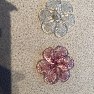 May include: Two clear, flower-shaped glass embellishments. One is transparent, the other is a soft pink. Each flower has six petals and a textured surface, creating a sparkling effect. These decorative elements could be used for crafts or jewelry.
