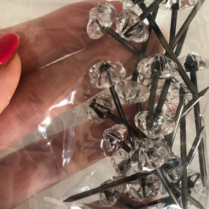May include: A close-up of a bag of clear glass tacks with black metal tips. The tacks are in a clear plastic bag and are being held by a hand.