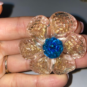 Puede incluir: Una flor de vidrio transparente con motas doradas y un centro azul. La flor tiene un diámetro de aproximadamente 5 cm.