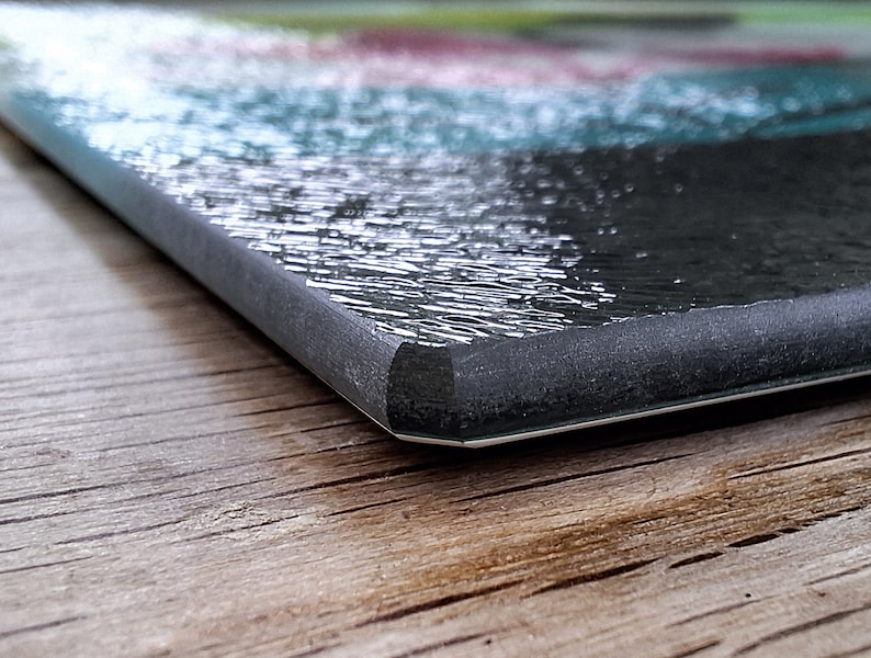 May include: Close-up of the edge of a black glass cutting board with a textured surface. The board is sitting on a wooden surface.