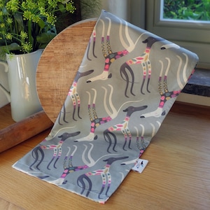 May include: A grey tea towel with a repeating pattern of stylised greyhounds with colourful geometric patterns. The towel is folded and lying on a wooden surface.