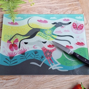 May include: A glass cutting board with a colorful design featuring two stylized greyhounds running through a field of flowers and butterflies. The board has a light blue background with pink, yellow, and green accents.