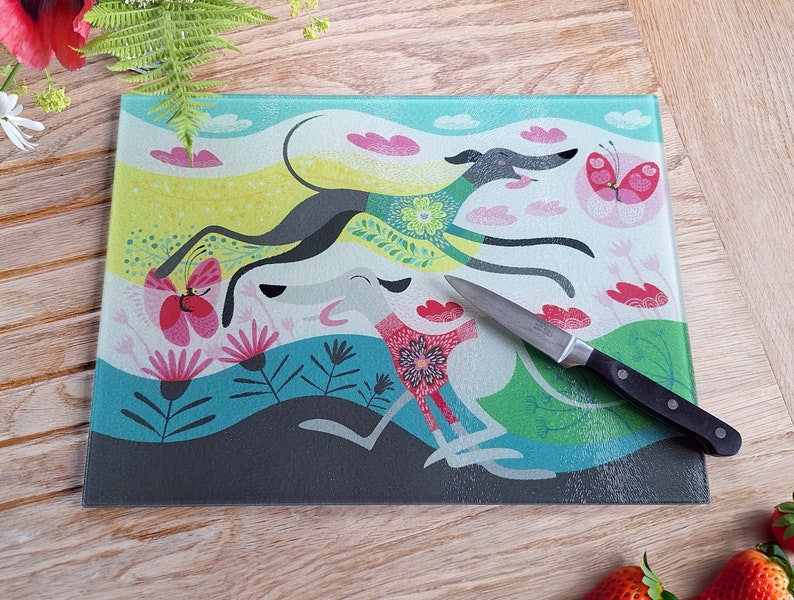 May include: A glass cutting board with a colorful design featuring two stylized greyhounds running through a field of flowers and butterflies. The board has a light blue background with pink, yellow, and green accents.