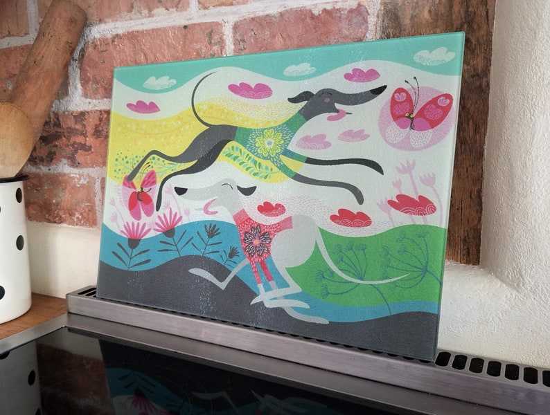 May include: A glass cutting board with a colorful illustration of three greyhounds running through a field of flowers and butterflies. The background is a light blue with pink clouds.