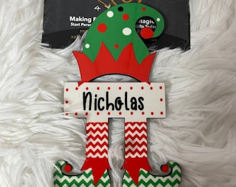 May include: A wooden elf ornament with a red and white chevron pattern on the legs, a green hat with red and white polka dots, and the name "Nicholas" written on the front.
