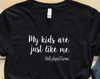 May include: Black v-neck t-shirt with the text "My kids are just like me - Well played Karma". The shirt is displayed on a wooden surface with a pair of jeans, sunglasses, and white sneakers.