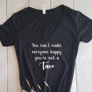 May include: Black V-neck t-shirt with the text "You can't make everyone happy, you're not a Taco" in white script. The shirt is decorated with a sprig of purple flowers and green leaves.