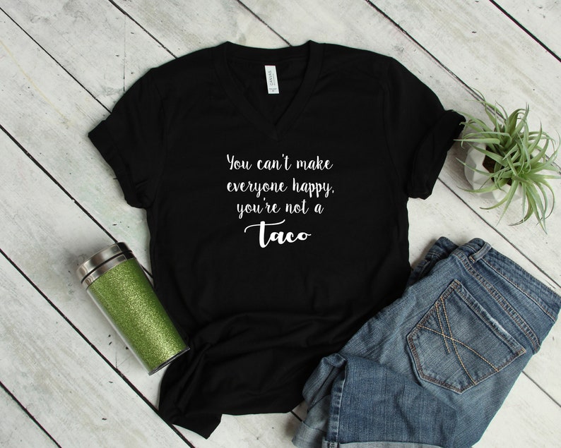 May include: Black V-neck t-shirt with the white text "You can't make everyone happy, you're not a Taco." The shirt is paired with blue denim shorts. A green glitter tumbler and a small potted plant are also in the image.