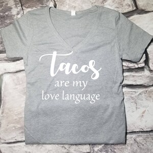 以下が含まれることがあります： 白い文字で「tacos are my love language」と書かれたグレーのVネックTシャツ
