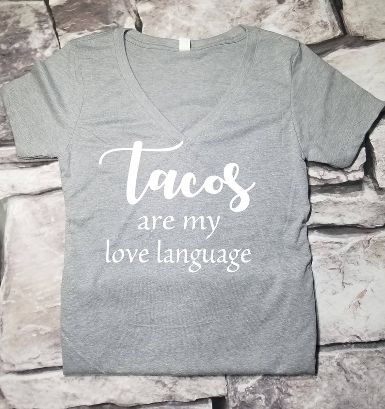 以下が含まれることがあります： 白い文字で「tacos are my love language」と書かれたグレーのVネックTシャツ
