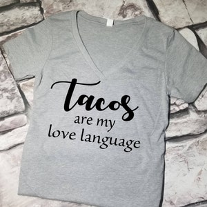 以下が含まれることがあります： グレーのVネックTシャツに「Tacos are my love language」という黒い文字がプリントされています。