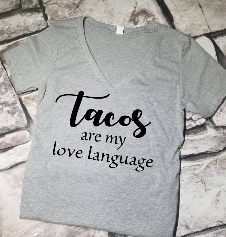 以下が含まれることがあります： グレーのVネックTシャツに「Tacos are my love language」という黒い文字がプリントされています。