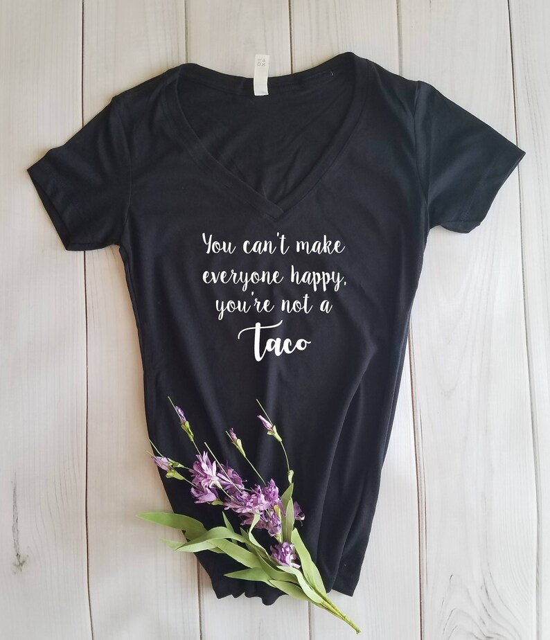 May include: Black V-neck t-shirt with the text "You can't make everyone happy, you're not a Taco" in white script. The shirt is decorated with a sprig of purple flowers and green leaves.