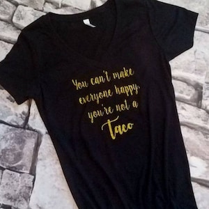 May include: Black V-neck t-shirt with gold glitter text. The text reads "You can't make everyone happy you're not a Taco". The shirt is made of a soft material and is suitable for casual wear.