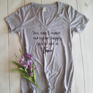 May include: A heather gray V-neck t-shirt with the text "You can't make everyone happy, you're not a Taco" in black script. A sprig of purple flowers rests on the shirt. The shirt is made of a soft material.