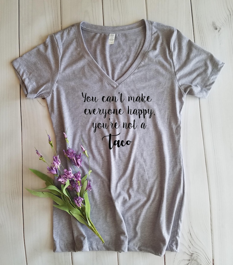May include: A heather gray V-neck t-shirt with the text "You can't make everyone happy, you're not a Taco" in black script. A sprig of purple flowers rests on the shirt. The shirt is made of a soft material.