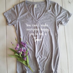 May include: A heather gray V-neck t-shirt with the text "You can't make everyone happy, you're not a Taco" in white script. A small bouquet of purple flowers with green leaves rests on the shirt. The shirt is on a white wooden surface.