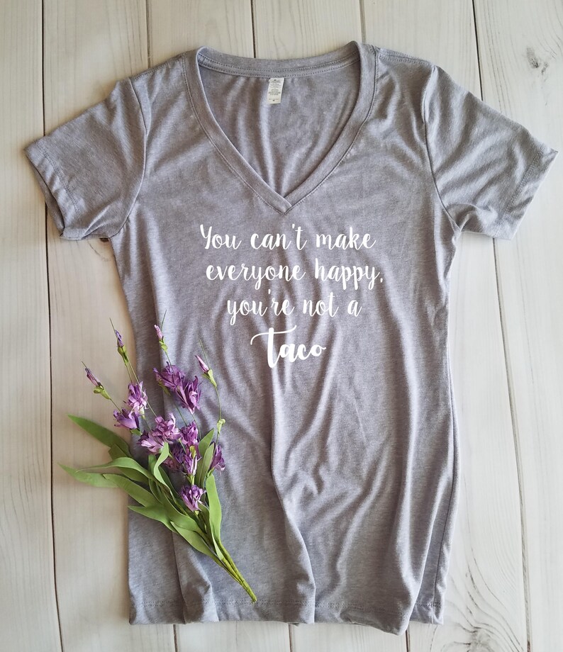 May include: A heather gray V-neck t-shirt with the text "You can't make everyone happy, you're not a Taco" in white script. A small bouquet of purple flowers with green leaves rests on the shirt. The shirt is on a white wooden surface.